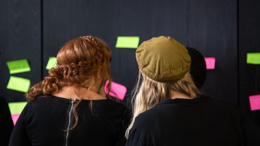 Kaksi naista katsovat mustaa seinää, johon on kiinnitetty värillisiä muistilappuja.