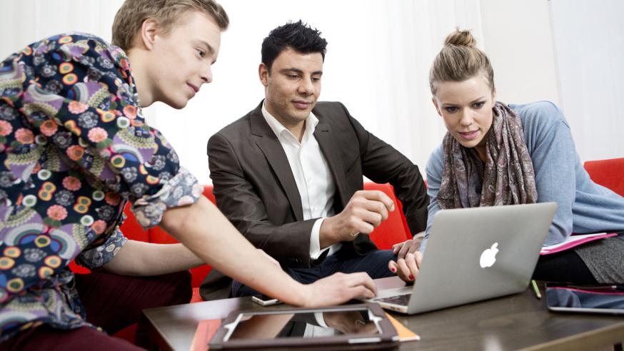 Three young adults sitting together focused on a laptop in front of them.