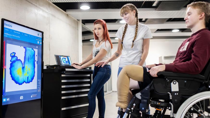 Kaksi apuvälinetekniikan opiskelijaa katsoo pyörätuolissa olevan asiakkaan kanssa näyttöä.