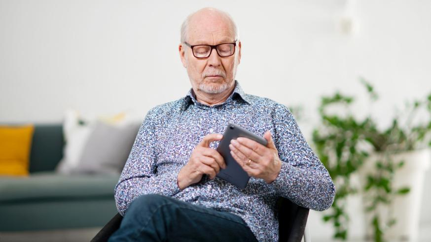 Ikääntynyt mies istuu ja tutkii kädessään olevaa mobiililaitetta.