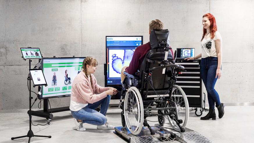 Apuvälinetekniikan opiskelijat säätävät opetusasiakkaan pyörätuolia.